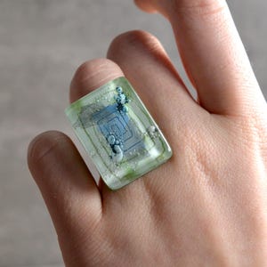 Op de afbeelding: Een rechthoekige ring met een doorschijnende lichtgroene basis en een centraal blauw vierkant spiraalpatroon, geaccentueerd door kleine witte belletjes en zilverkleurige metaalschilfers ingebed in het gladde, glanzende oppervlak van het materiaal.