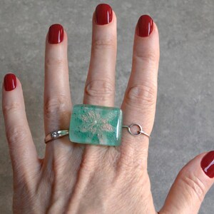 Può includere: Un anello caratterizzato da un elemento centrale rettangolare in vetro traslucido color verde acqua con un motivo floreale bianco incorporato. Il pezzo in vetro è montato su una sottile fascia in metallo argentato che avvolge il dito, arricchito da un piccolo dettaglio circolare.