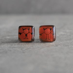 May include: A pair of square stud earrings. Each earring has a red background with a black cartoon devil illustration. The devil has horns, arms, and legs. The earrings have a glossy finish.