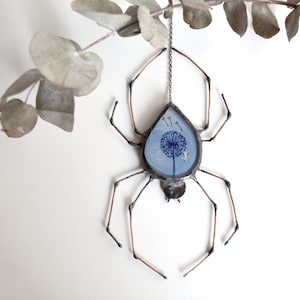 Puede incluir: Una araña de vidrio azul con un diseño de diente de león colgando de una cadena. La araña está hecha de alambre de cobre y tiene un aspecto delicado y caprichoso.