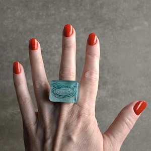 May include: A square, turquoise ring with a water-like design is worn on a hand with red nail polish. The ring is made of a translucent material, and the design features concentric circles. The background is a neutral grey.