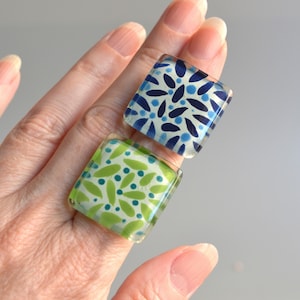 May include: Two square rings with smooth, glossy surfaces. One ring features a pattern of dark blue petals and small light blue dots on a pale background. The other ring displays a pattern of lime green petals and dark teal dots on a pale background.