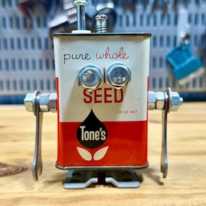 May include: A handcrafted robot sculpture made from a repurposed Tone's seed can. The robot features bolt eyes, metal legs, and a silver antenna. The can is white and red with the words "pure whole SEED" and "Tone's" printed on it. The net weight is 1 ½ oz.