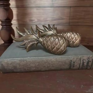 May include: Two metallic, brass-toned pineapple-shaped decorative objects featuring textured, cross-hatched bodies and pointed, leafy tops. These items are positioned horizontally, resting on a thick, grey-covered book with visible text on the spine.