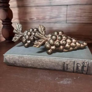 May include: A pair of metallic gold-toned grape clusters with detailed leaves and stems resting on top of a rectangular book titled Another Claudia by Rose Franken. The grapes feature a textured, rounded surface with a matte finish.