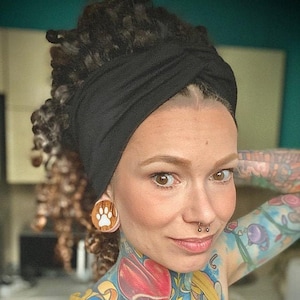 Op de afbeelding: Een vrouw met zwarte dreadlocks en kleurrijke tatoeages draagt een zwarte turban en een paar houten oorpluggen met een pootprintontwerp. Ze draagt een geel tanktop.