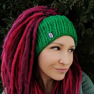 May include: A woman with long, burgundy dreadlocks is wearing a green knitted headband with a small skull design. The headband is worn above her forehead and covers her ears.