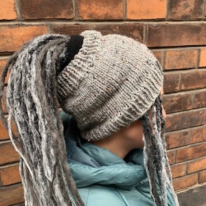 May include: A grey knitted beanie with a black ponytail holder. The beanie is being worn by a person with long grey and black dreadlocks.