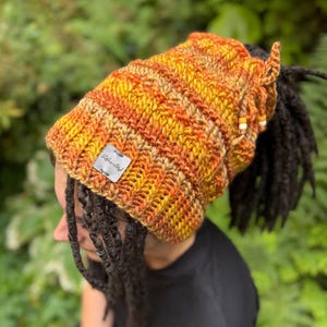 May include: A crocheted hat in shades of orange, yellow, and brown, with a tag that reads "Afro-Styl". The hat is designed to accommodate dreadlocks, with a ponytail opening at the back. The hat is adorned with small beads.