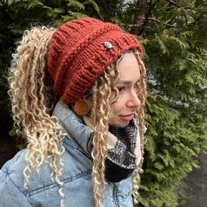 Könnte beinhalten: Eine rostige Strickmütze mit einem Pferdeschwanzloch und einem silbernen Knopf oben. Die Mütze wird von einer Person mit blonden Dreadlocks und einer Jeansjacke getragen.