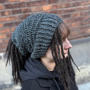 May include: A gray crocheted beanie with a textured pattern. The beanie is worn with dark dreadlocks. The person is wearing a black jacket with a hood. The background is a brick wall.