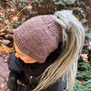 May include: A brown knitted beanie with a ponytail hole. The beanie has a small brown leather tag with the word "Handmade" on it. The beanie is being worn by a person with long, gray dreadlocks.