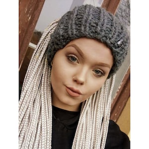 以下が含まれることがあります： 白とグレーの長い編み込みの髪をした女性が、黒いロゴの付いたグレーのニット帽をかぶっています。