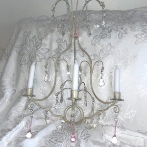 May include: A wall-mounted candle holder with a light beige metal frame featuring three candle cups, each holding a white taper candle. The structure is adorned with clear and pink glass teardrop crystals hanging from curved metal arms.