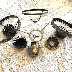 May include: A collection of disassembled lamp parts including two circular metal frames with ornate filigree edges, a central brass burner assembly with a small key, a circular support ring, and two additional brass-toned circular components.