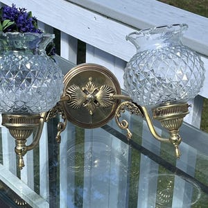 May include: A vintage double sconce with clear, diamond-patterned glass shades and ornate brass fixtures. The sconce features a central decorative plate and curved arms, adding a touch of elegance. The sconce is designed to be mounted on a wall.