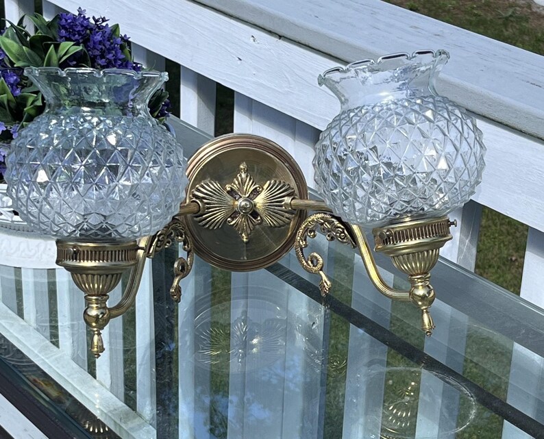 May include: A vintage double sconce with clear, diamond-patterned glass shades and ornate brass fixtures. The sconce features a central decorative plate and curved arms, adding a touch of elegance. The sconce is designed to be mounted on a wall.