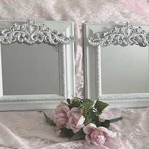 May include: Two square mirrors with white frames featuring ornate, carved scrollwork crests at the top. The frames have a distressed finish with visible texture. A small bouquet of light pink artificial roses is positioned in front of the mirrors.