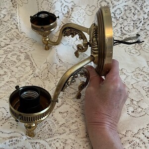 May include: Ornate brass wall sconce with two light fixtures. The sconce features decorative scrollwork and a circular base. The light fixtures have a dark interior and are designed to hold light bulbs. Electrical wires are visible.