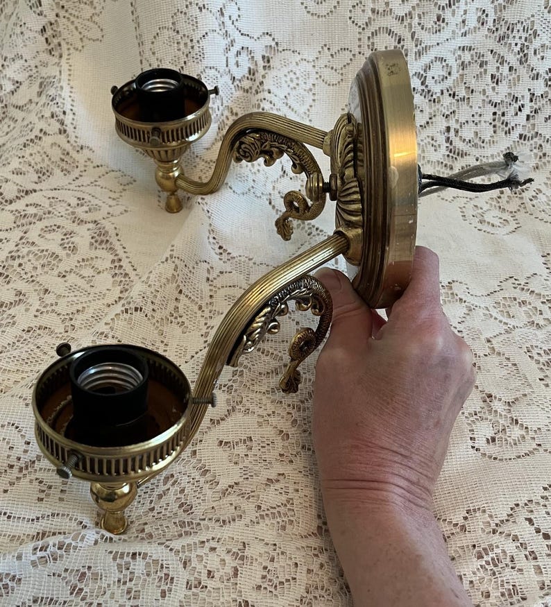 May include: Ornate brass wall sconce with two light fixtures. The sconce features decorative scrollwork and a circular base. The light fixtures have a dark interior and are designed to hold light bulbs. Electrical wires are visible.