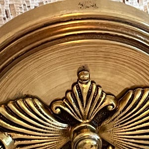May include: Close-up of a gold-colored, ornate decorative object. The object features a central, circular design with a detailed, fan-like pattern radiating outwards. The surface has a textured, aged appearance, with multiple concentric rings.