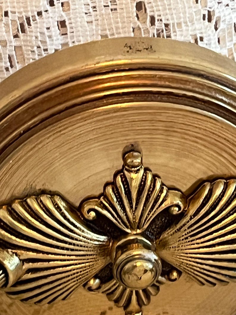 May include: Close-up of a gold-colored, ornate decorative object. The object features a central, circular design with a detailed, fan-like pattern radiating outwards. The surface has a textured, aged appearance, with multiple concentric rings.