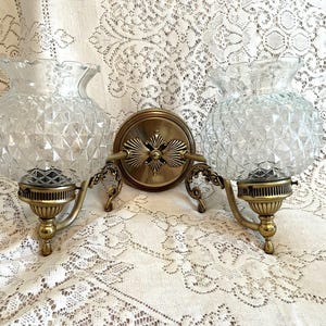 May include: A vintage double sconce with clear, diamond-patterned glass shades and ornate brass fixtures. The sconce features a central circular mounting plate and decorative arms supporting the glass globes. The design evokes a classic, elegant aesthetic.