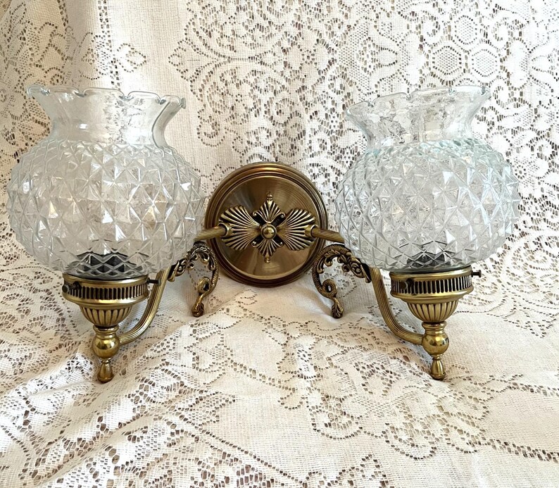 May include: A vintage double sconce with clear, diamond-patterned glass shades and ornate brass fixtures. The sconce features a central circular mounting plate and decorative arms supporting the glass globes. The design evokes a classic, elegant aesthetic.