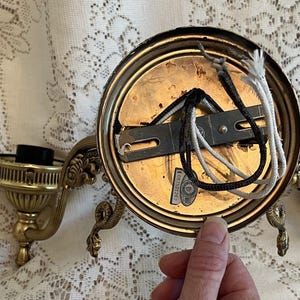 May include: Vintage brass wall sconce with ornate detailing. The sconce features a circular backplate, two candle holders, and decorative arms. Electrical wiring is visible. A hand is holding the sconce.