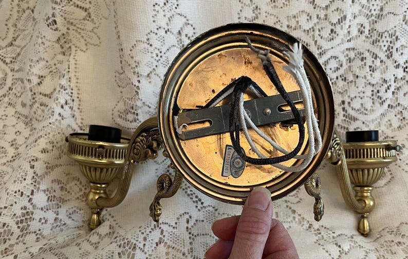 May include: Vintage brass wall sconce with ornate detailing. The sconce features a circular backplate, two candle holders, and decorative arms. Electrical wiring is visible. A hand is holding the sconce.