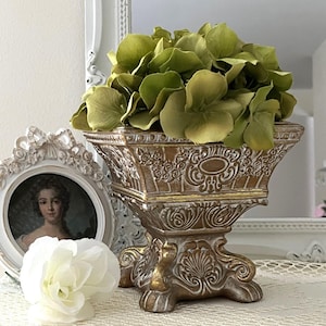 May include: A decorative, ornate gold and bronze-colored pedestal vase filled with green faux flowers. The vase has a detailed, embossed design and sits on a decorative base. A framed portrait and a white flower are in the background.