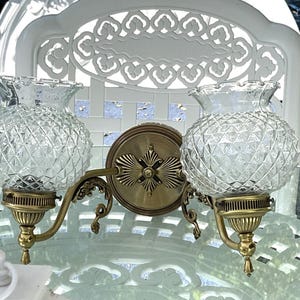 May include: A vintage double sconce with clear, diamond-patterned glass globes and brass-toned metal fixtures. The sconce features a decorative backplate and curved arms, adding a touch of classic elegance to any space.