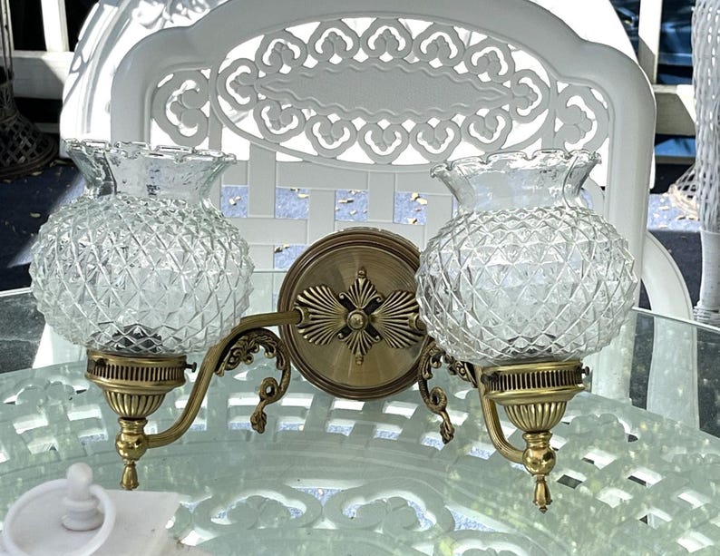 May include: A vintage double sconce with clear, diamond-patterned glass globes and brass-toned metal fixtures. The sconce features a decorative backplate and curved arms, adding a touch of classic elegance to any space.