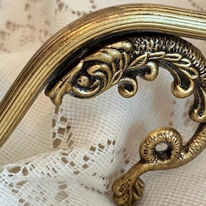 May include: Close-up of a gold-colored, ornate decorative element. The piece features a curved, ribbed bar and a detailed, scroll-like design with a textured finish. The decorative element is likely part of a larger vintage furniture piece.