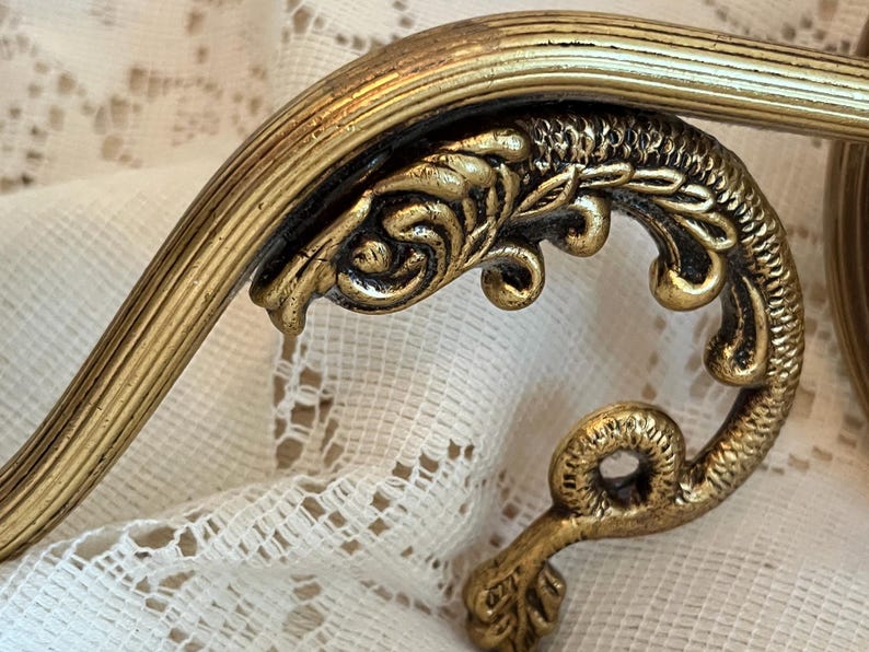 May include: Close-up of a gold-colored, ornate decorative element. The piece features a curved, ribbed bar and a detailed, scroll-like design with a textured finish. The decorative element is likely part of a larger vintage furniture piece.
