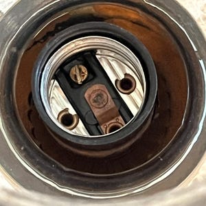 May include: Close-up of a light fixture socket. The socket has a black interior with copper-colored contacts and a silver-colored metal rim. The socket is designed to hold a light bulb.