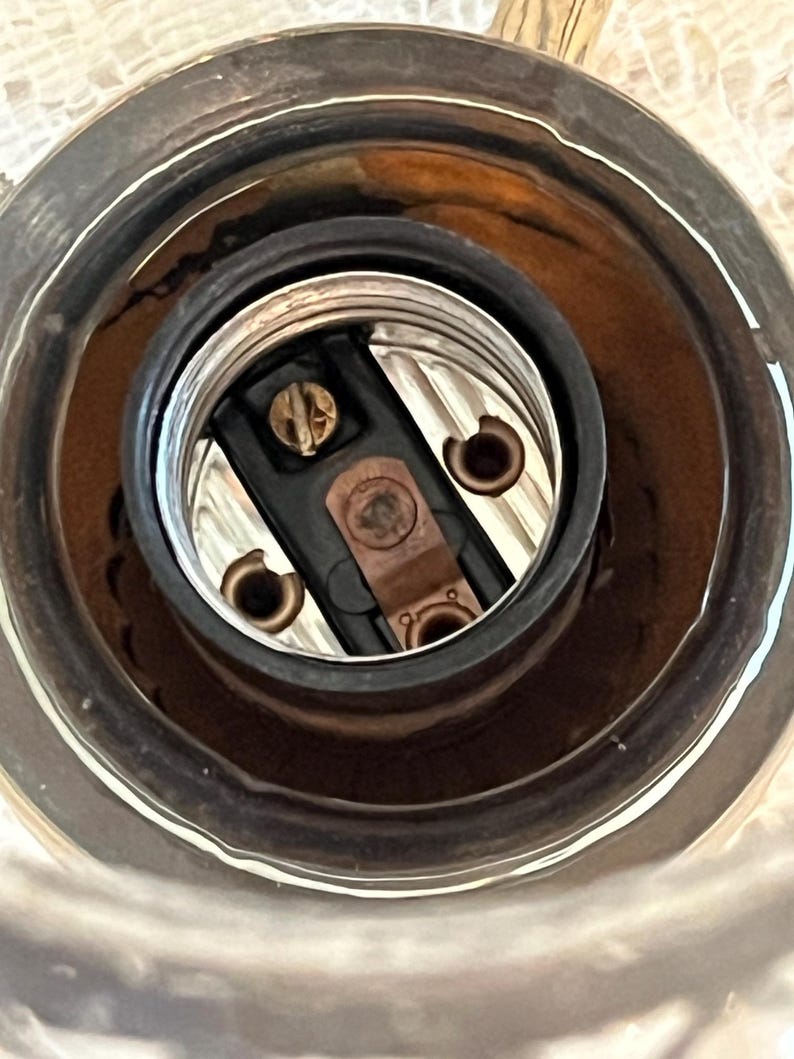 May include: Close-up of a light fixture socket. The socket has a black interior with copper-colored contacts and a silver-colored metal rim. The socket is designed to hold a light bulb.