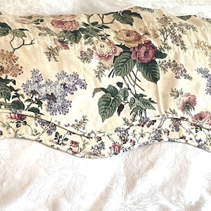 May include: A window valance featuring a cream background with a floral pattern of pink roses, purple lilacs, and green leaves. The fabric has a scalloped bottom edge with a matching floral trim, creating a decorative border along the lower hem.