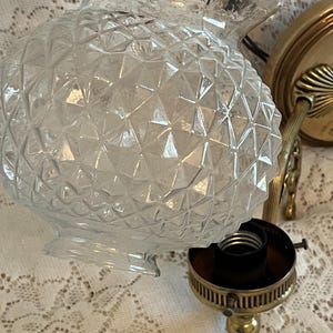 May include: Clear glass light fixture globe with a diamond pattern. The globe has a ruffled edge and is held near a brass-colored light fixture base. The base includes a bulb socket and a decorative brass arm.