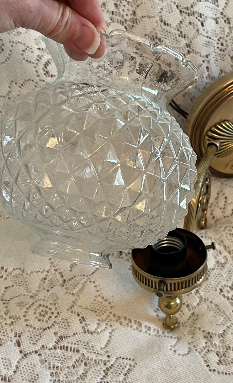 May include: Clear glass light fixture globe with a diamond pattern. The globe has a ruffled edge and is held near a brass-colored light fixture base. The base includes a bulb socket and a decorative brass arm.