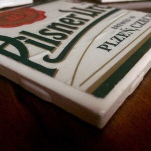 K&ouml;nnte beinhalten: Wei&szlig;er Keramikuntersetzer mit dem Pilsner Urquell Logo und dem Text "Brewed in Plzen, Czech Republic".