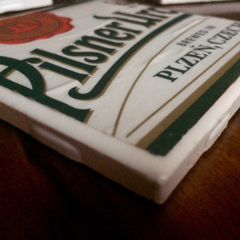 K&ouml;nnte beinhalten: Wei&szlig;er Keramikuntersetzer mit dem Pilsner Urquell Logo und dem Text "Brewed in Plzen, Czech Republic".