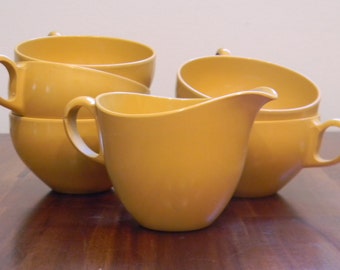 May include: Set of four yellow ceramic mugs and a yellow ceramic creamer. The mugs have a rounded shape and a handle on the side. The creamer has a rounded shape with a handle on the side and a spout on the front.