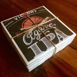 K&ouml;nnte beinhalten: Ein Stapel von sechs schwarzen und wei&szlig;en Keramik-Untersetzern mit braunem Hintergrund. Die Untersetzer zeigen den Text "Victory Brewing Company" und "Agave IPA" mit einer Grafik einer Orangenscheibe. Die Untersetzer sind mit Schnur zusammengebunden.
