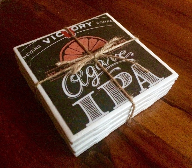 K&ouml;nnte beinhalten: Ein Stapel von sechs schwarzen und wei&szlig;en Keramik-Untersetzern mit braunem Hintergrund. Die Untersetzer zeigen den Text "Victory Brewing Company" und "Agave IPA" mit einer Grafik einer Orangenscheibe. Die Untersetzer sind mit Schnur zusammengebunden.