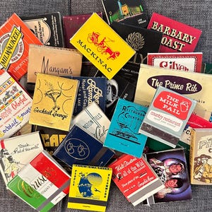 May include: A collection of vintage matchbook covers with various designs and text. The covers feature images of cocktails, restaurants, hotels, and other businesses. Some of the text includes "The Prime Rib", "The Milk Pail", "Barbary Coast", and "Cocktail Lounge".