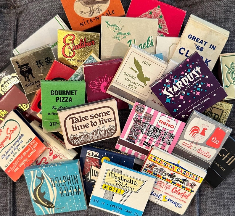May include: A collection of vintage matchboxes with various designs and colors. Some of the matchboxes feature text, including "Nite-N-Sale", "Gourmet Pizza", "Join Ducks Unlimited Now", "Great In 768", "Stardust Hotel and Casino", "Take some time to live", "Circus in Reno", "Cock-a-doodle", "Martinettis Dine Cocktails Motel", "Arlington Carousel", "Festive Colorful Tail Lounge", and "Chi Chicago".
