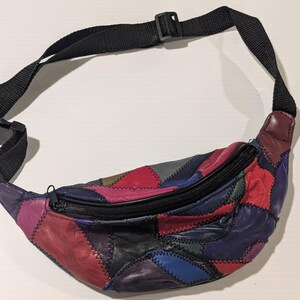 May include: A multicolored patchwork fanny pack constructed from various leather pieces in shades of red, blue, purple, and black. The bag features a black zippered closure across the top and an adjustable black nylon strap with a plastic buckle.