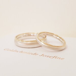 May include: Two wedding bands, one silver with a thin gold band around the edge, and the other silver with a thicker gold band around the edge. The bands are on a white surface with the text "Goldschmiede Josefine" printed in brown.