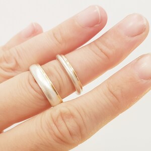 May include: Two wedding bands on a finger. The top band is a thin gold band with a white metal edge. The bottom band is a wider white metal band with a thin gold edge.